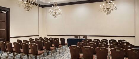 Un salón de conferencias con filas de sillas marrones, una mesa larga y dos candelabros.