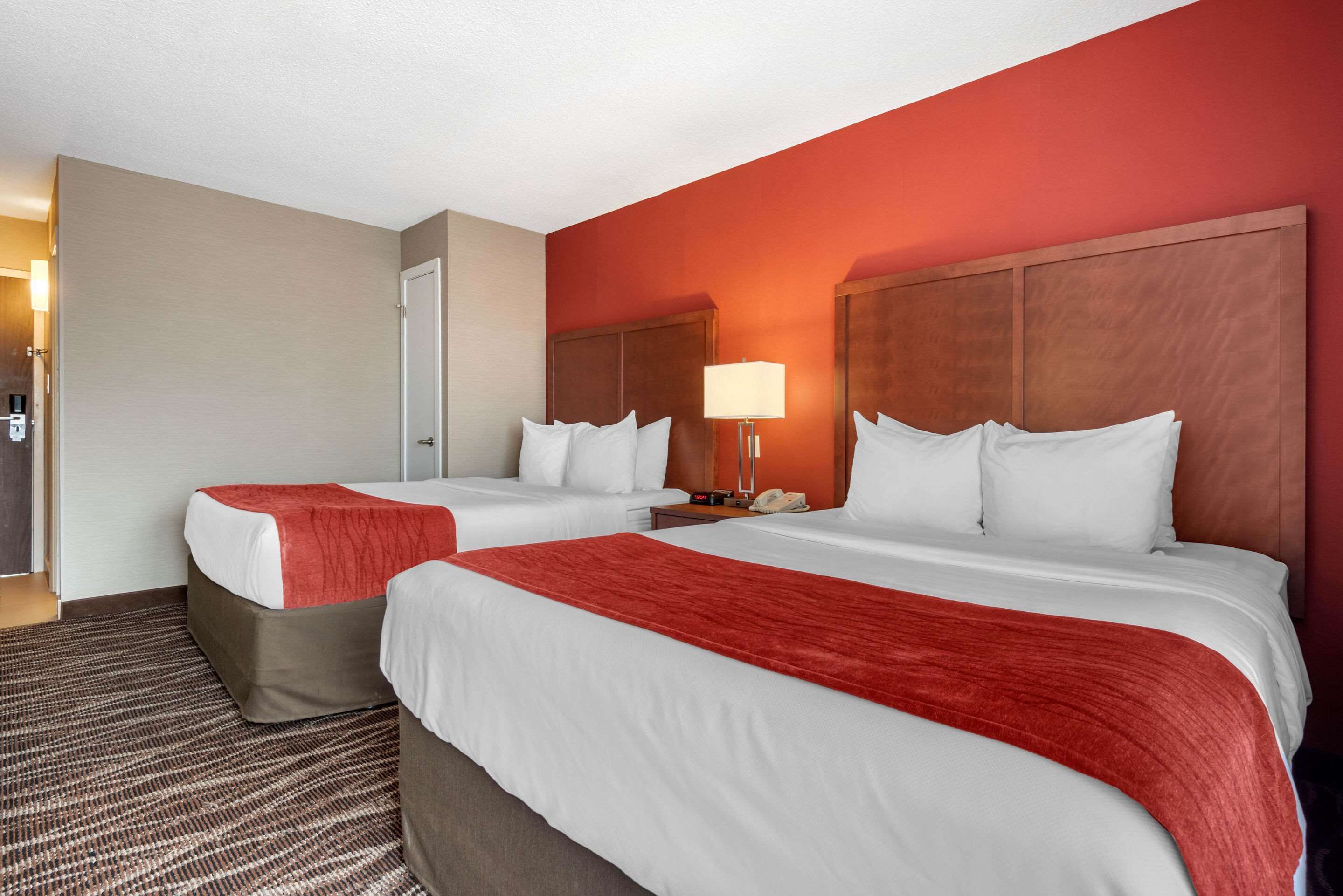 A hotel room with two beds, a red accent wall, a wooden headboard, a bedside table with a lamp, and a carpeted floor.