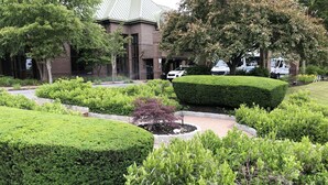 Ein liebevoll angelegter Garten mit ordentlich geschnittenen Hecken, einem kreisförmigen Blumenbeet in der Mitte und einem Backsteinhaus mit markanter Dachform im Hintergrund.