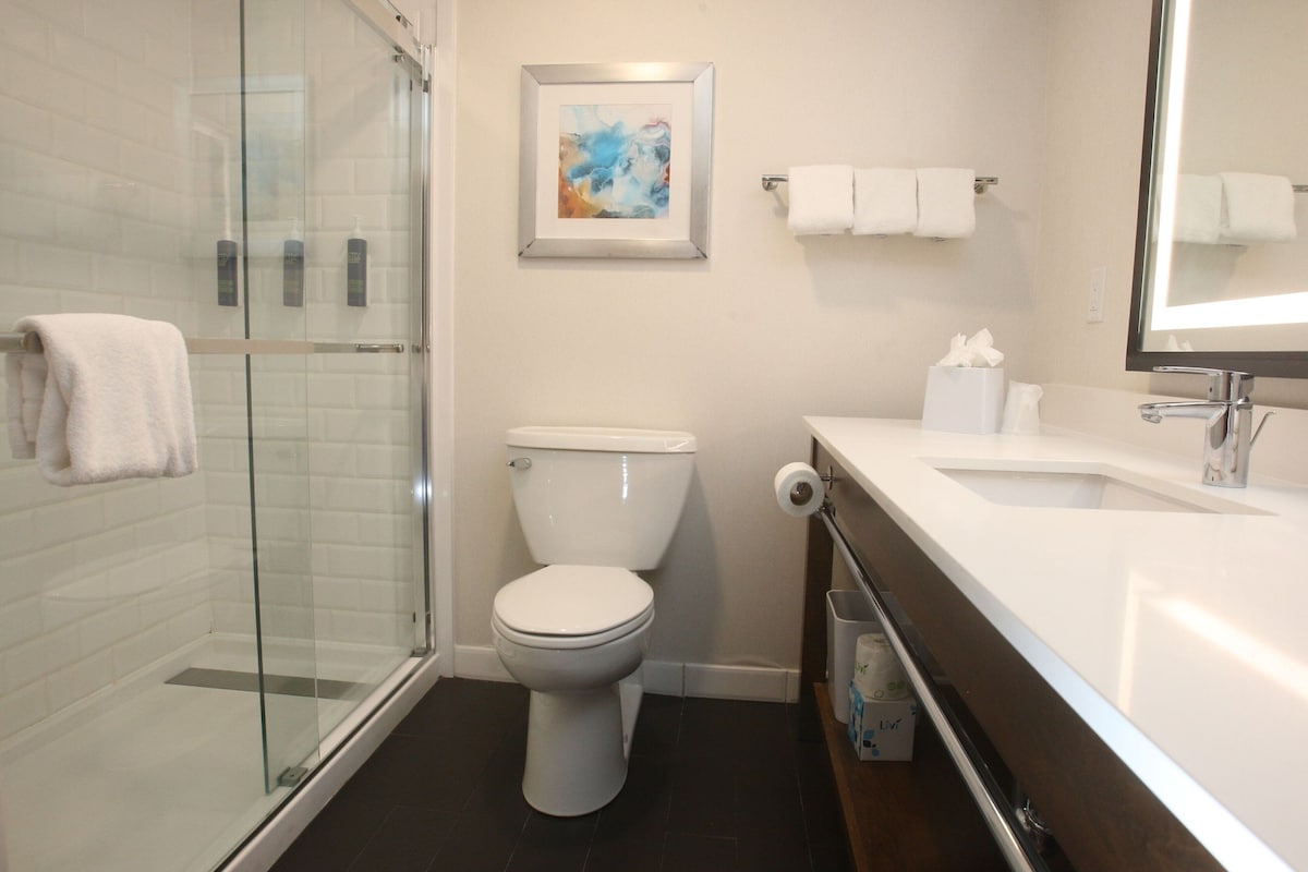 A modern bathroom with a glass shower enclosure, a toilet, a sink, and a framed abstract painting on the wall.