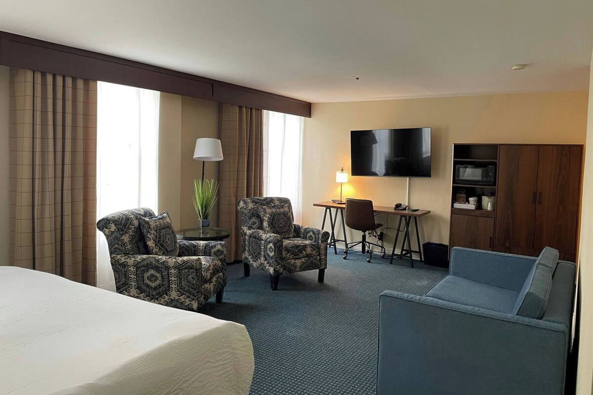 Ein Hotelzimmer mit einem Bett, zwei gemusterten Sesseln, einem Schreibtisch mit Lampe, einem Fernseher und einer Küchenzeile.