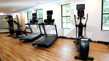 Een fitnessruimte met loopbanden, elliptische trainers en een hometrainer, met grote ramen en een houten vloer.