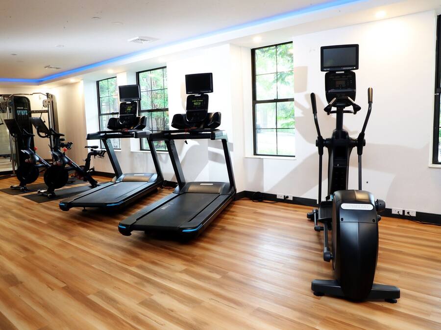 A gym with treadmills, elliptical machines, and a stationary bike, with large windows and a wooden floor.