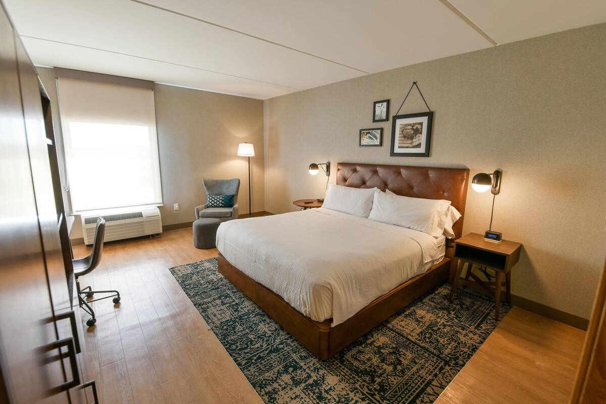 Ein ordentlich eingerichtetes Schlafzimmer mit einem großen Bett, einem Sessel, Nachttischen und einem Fenster mit Jalousien.