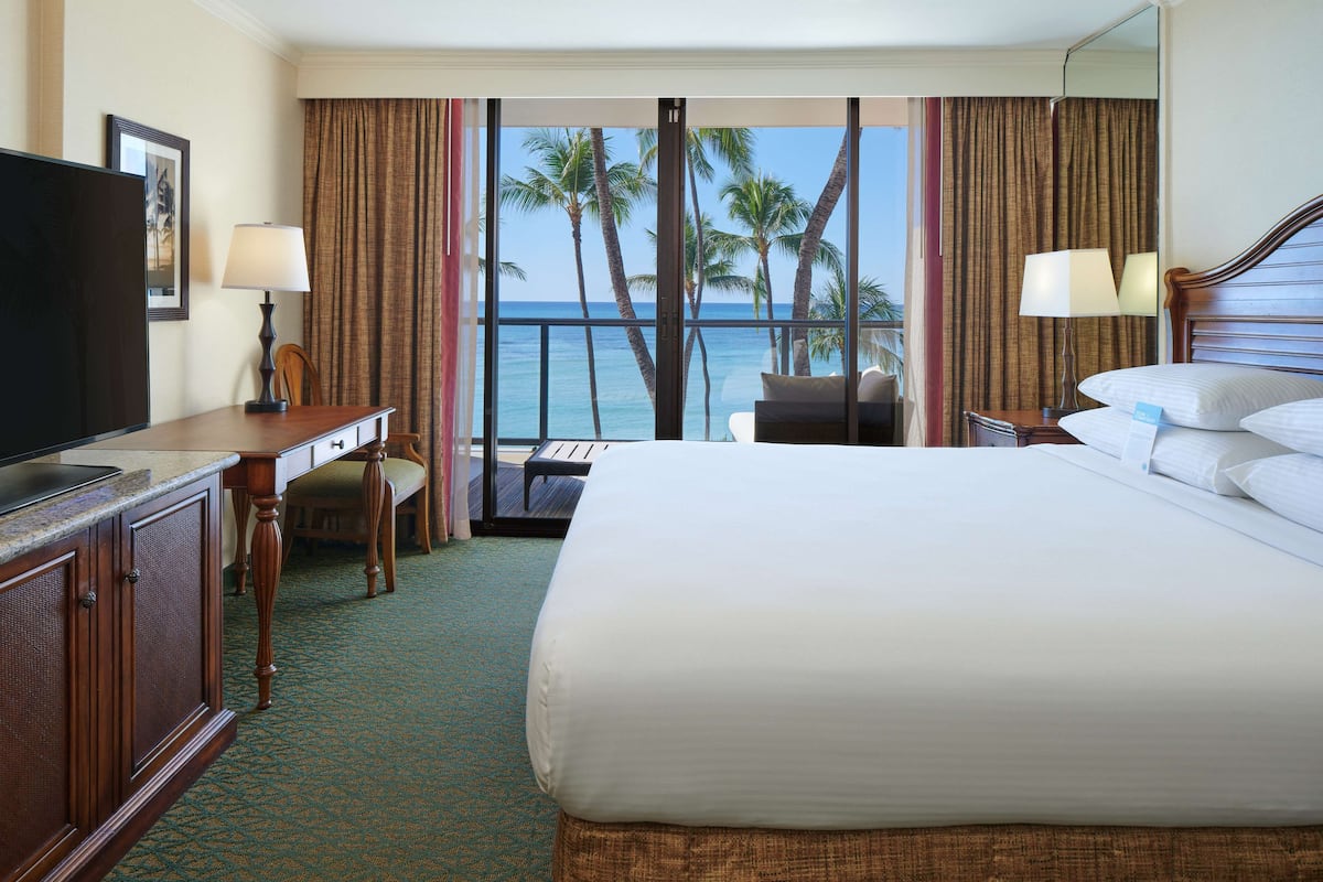A hotel room with a large bed, a desk, a television, and a view of palm trees and the ocean.