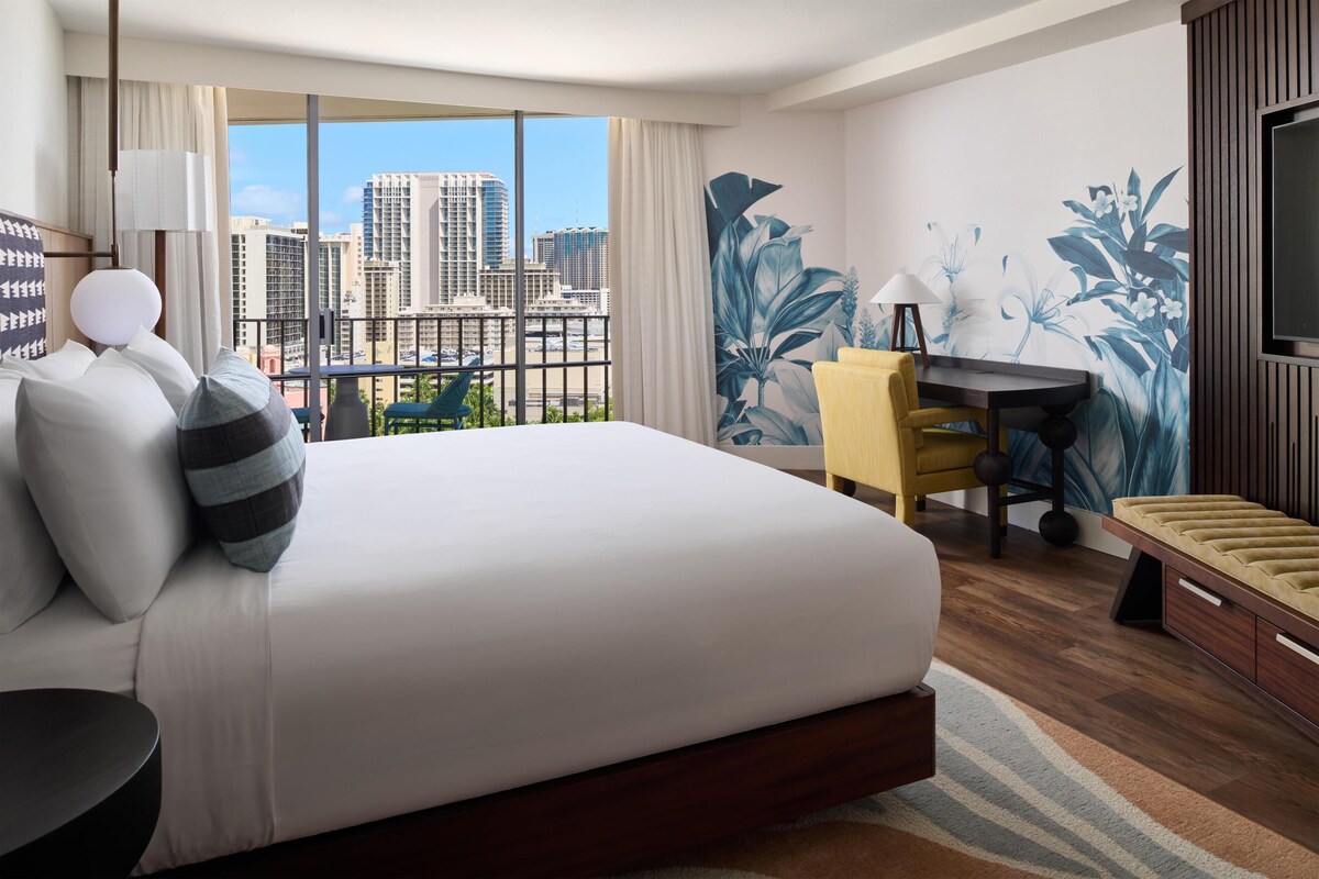 A hotel room with a large bed, a desk, a chair, a TV, and a city view.
