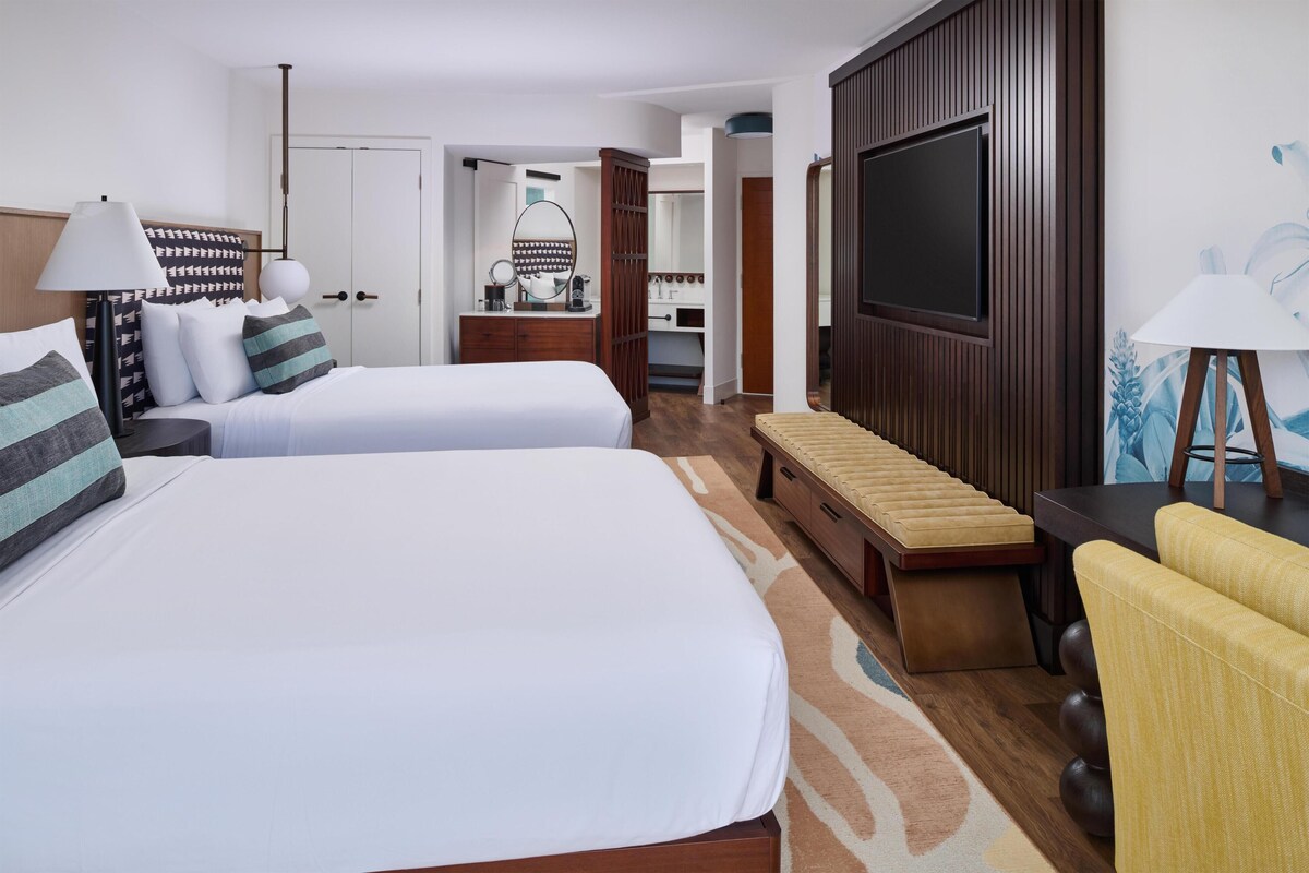 A hotel room with two beds, a TV, a bench, a chair, and a bathroom visible through an open door.