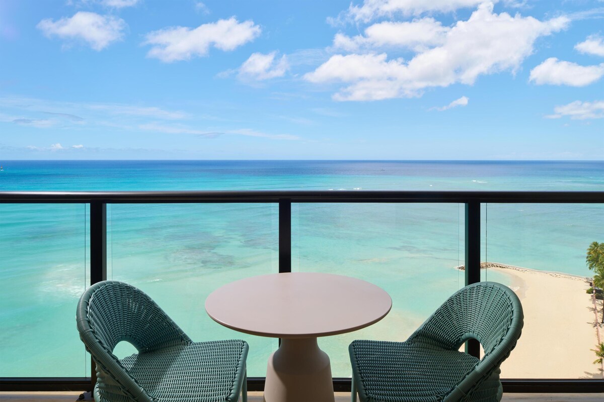 A balcony with two wicker chairs and a small table, overlooking a beach and turquoise sea.