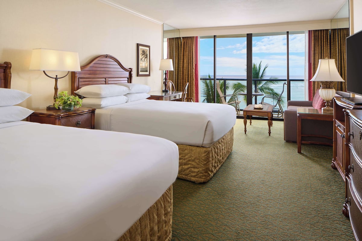 A hotel room with two beds, a desk, a chair, a TV, and a balcony with a view of the ocean.