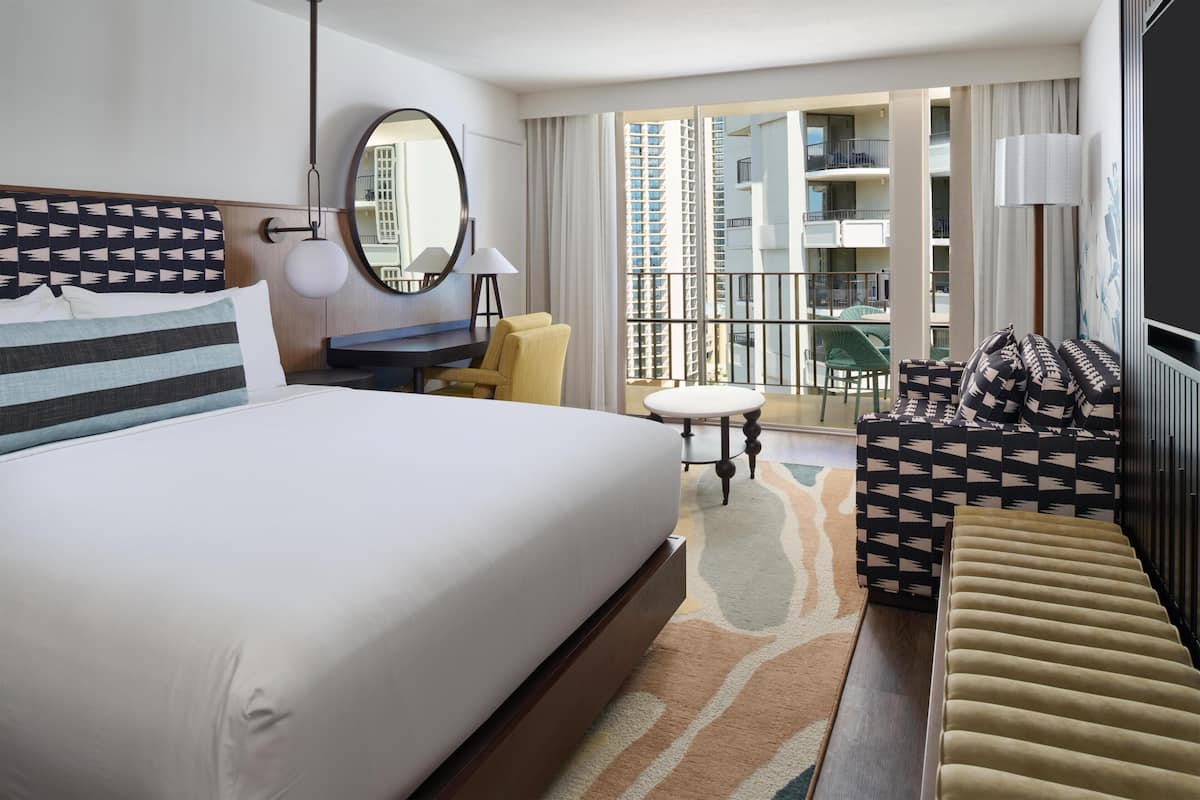 A hotel room with a large bed, a desk, a chair, a sofa, a round table, and a balcony with a view of high-rise buildings.