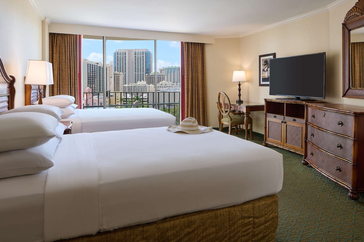A hotel room with two beds, a television, a desk with a chair, and a view of the cityscape.