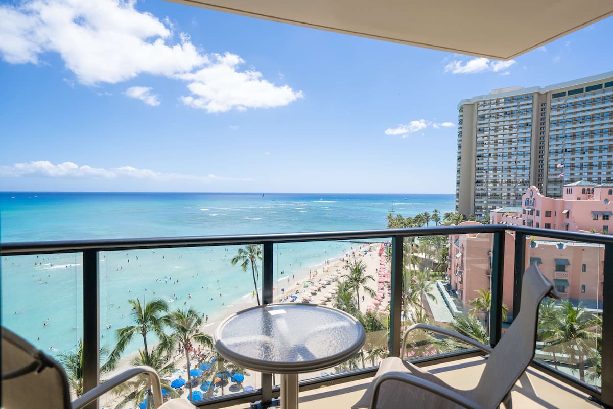 A balcony with a view of the beach, palm trees, and a hotel building.
