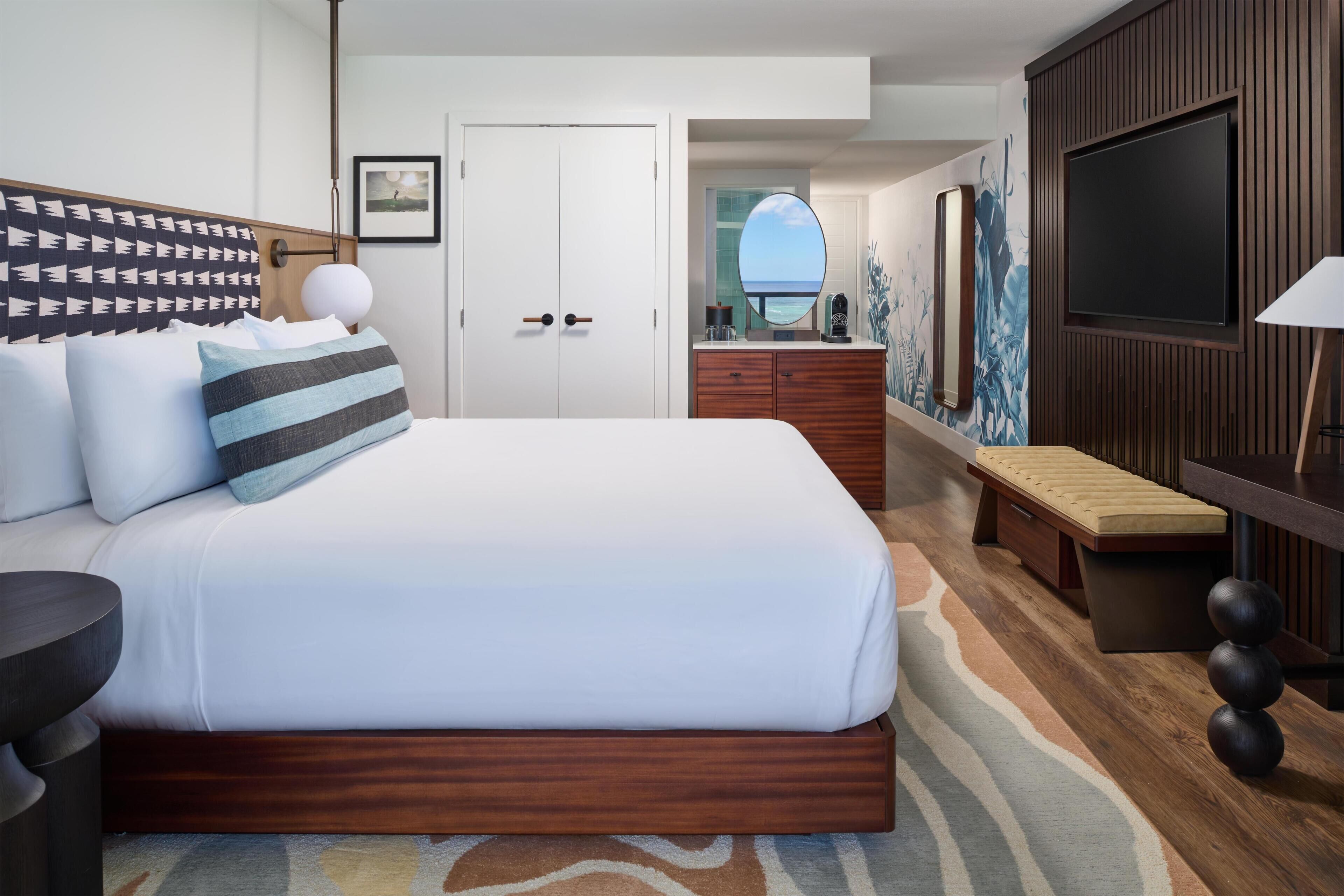 Een hotelkamer met een groot bed, een houten hoofdbord, een televisie, een bankje, een bureau en een spiegel.