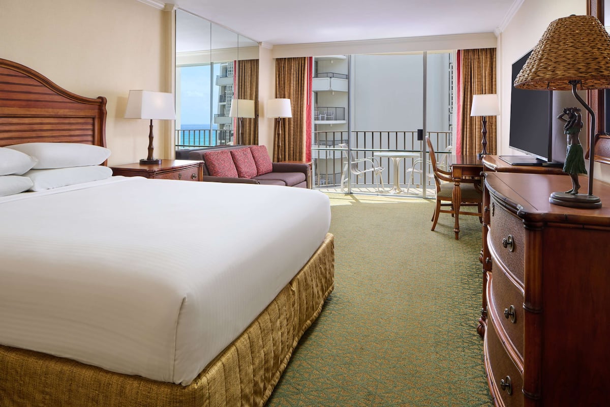 A hotel room with a large bed, a desk, a chair, a dresser, and a balcony with a view.