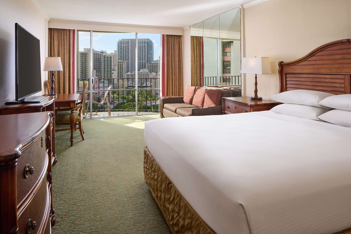 A hotel room with a large bed, a desk with a TV, a chair, a nightstand, and a view of the cityscape.