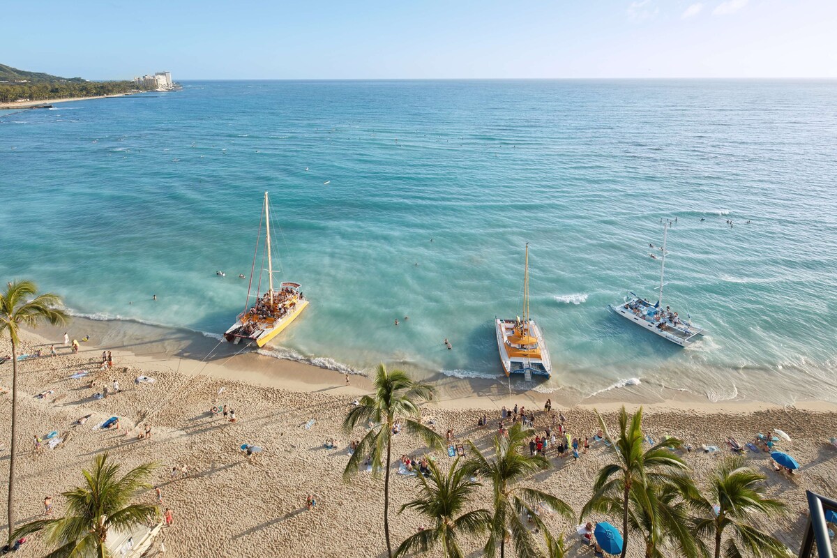 A beach with boats, palm trees, and people.