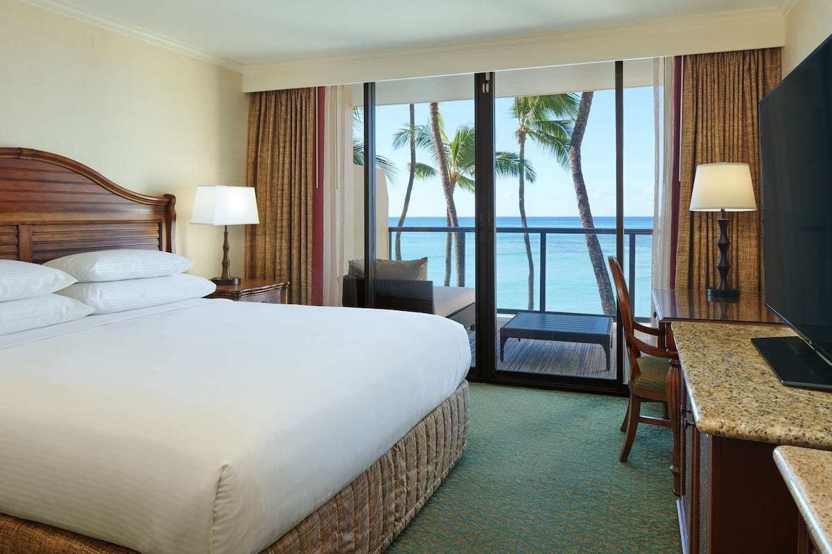 A hotel room with a bed, a desk, a chair, a TV, and a view of the ocean with palm trees.