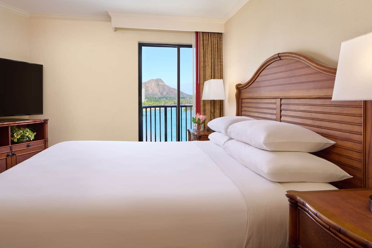 A hotel room with a large bed, a television, a nightstand, a lamp, and a view of a mountain through a balcony.