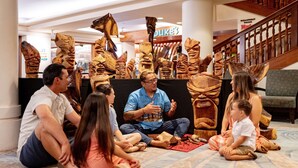 Un grupo de personas sentadas en el suelo alfombrado de un vestíbulo, con esculturas de madera al fondo.
