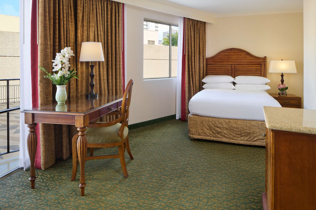 A hotel room with a bed, a desk with a chair, a window, and a nightstand with a lamp.