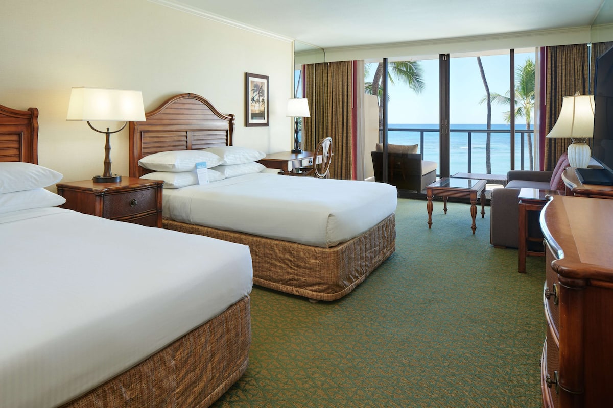 A hotel room with two beds, a desk, a chair, and a view of the ocean.