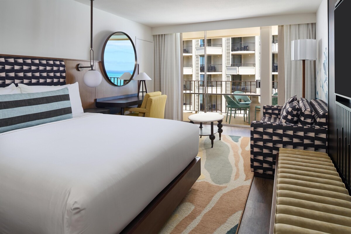 A hotel room with a large bed, a desk, a chair, a sofa, and a balcony with a view of the sea.