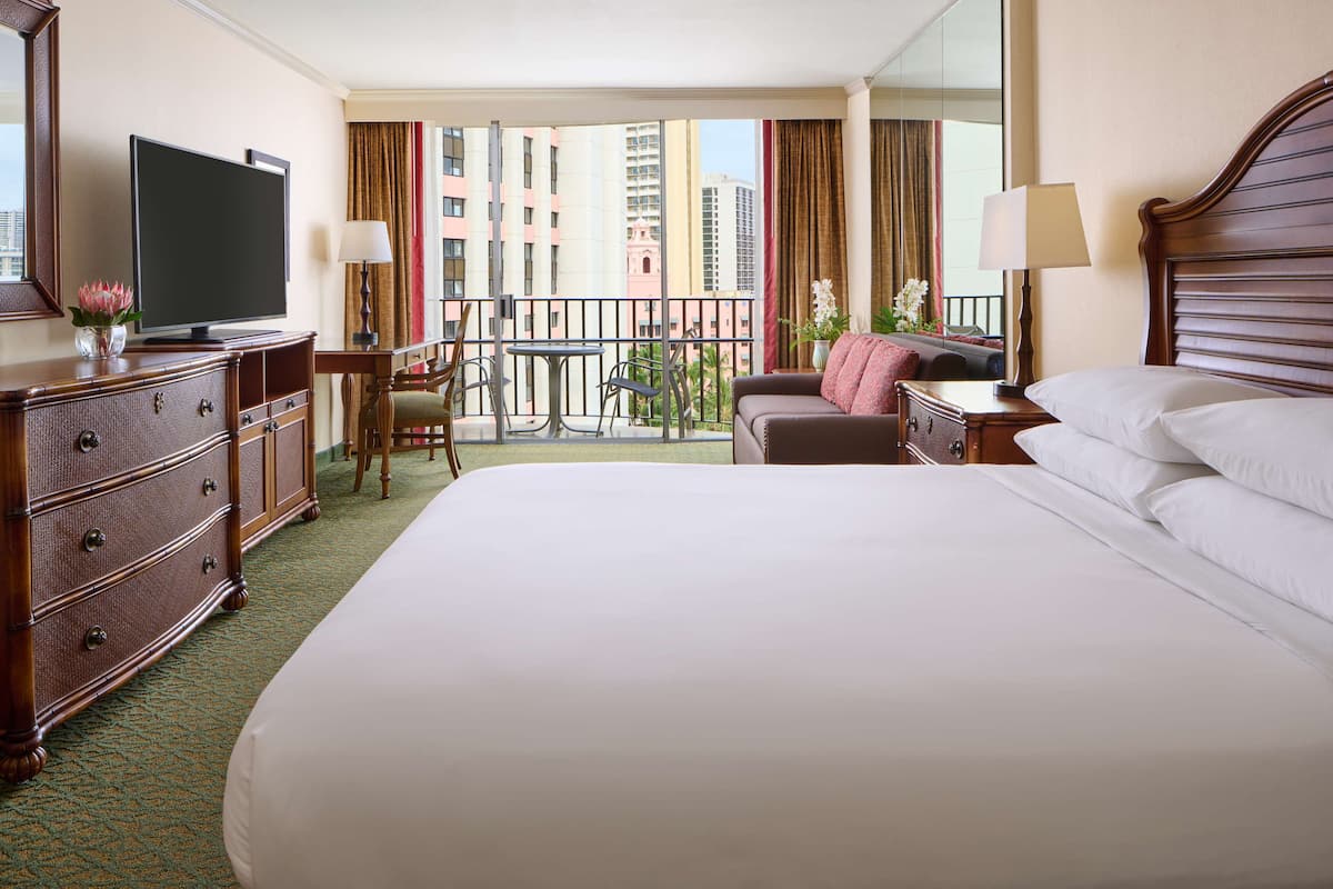 A hotel room with a large bed, a television, a desk with a lamp, a chair, and a balcony with a view of buildings.