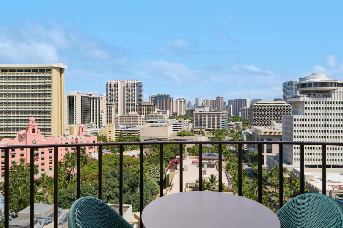 A balcony with a table and chairs, overlooking a cityscape with various buildings and palm trees.
