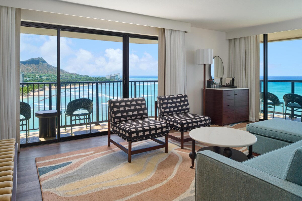 A hotel room with a balcony, two armchairs, a sofa, a small table, and a minibar.