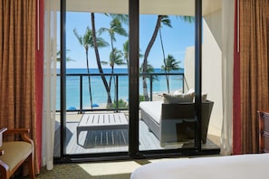 A hotel room with a balcony, a bed, a chair, and a view of the ocean and palm trees.