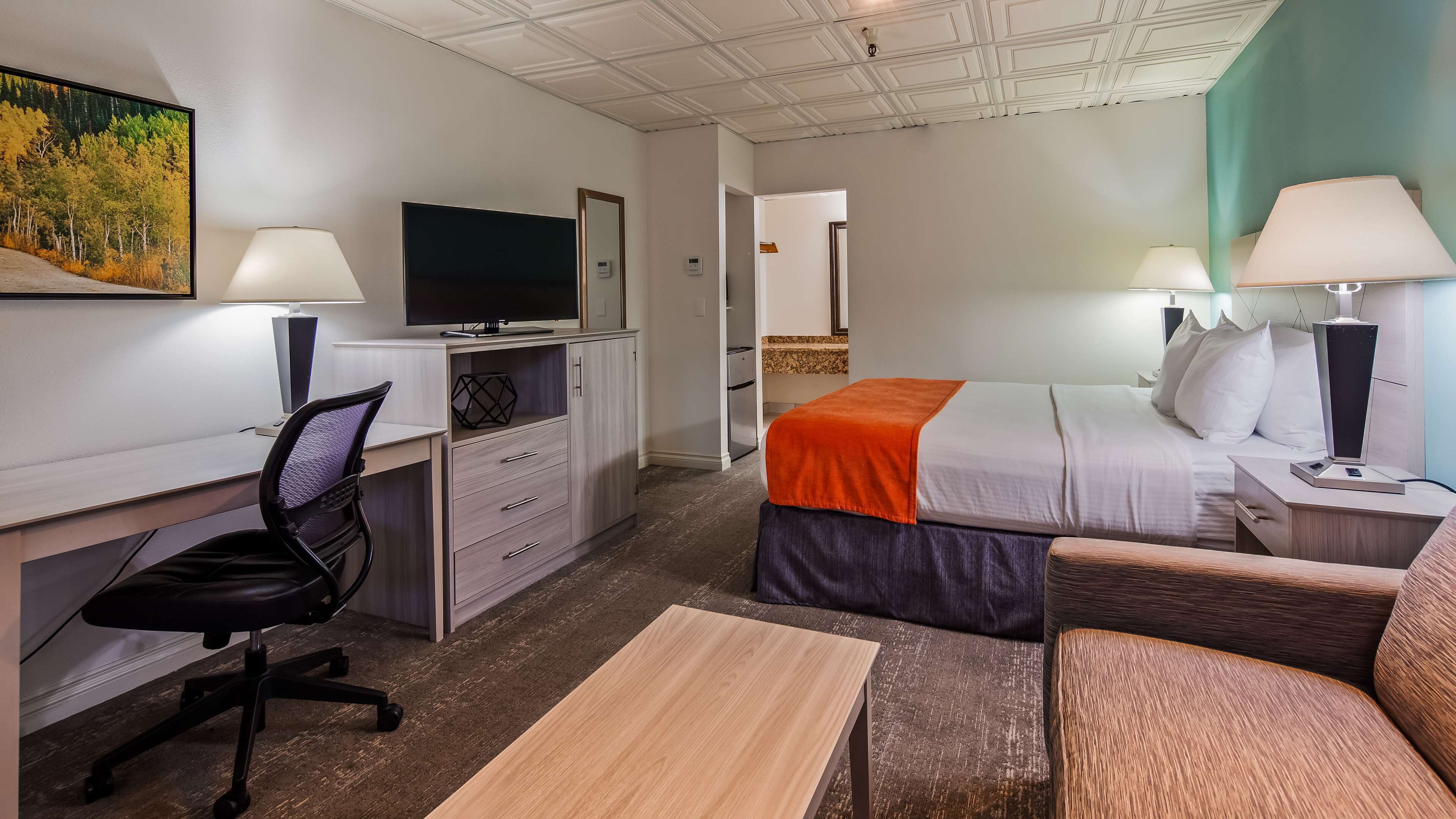 A hotel room with a desk, chair, bed, bedside tables, lamps, and a TV.