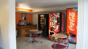 Una habitación pequeña e iluminada con dos mesas redondas y cuatro sillas, un microondas, un refrigerador y una máquina expendedora de Coca-Cola.
