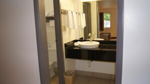 Un baño con encimera negra, lavamanos blanco y un espejo. Hay un perchero con toallas blancas y un cubo de basura.