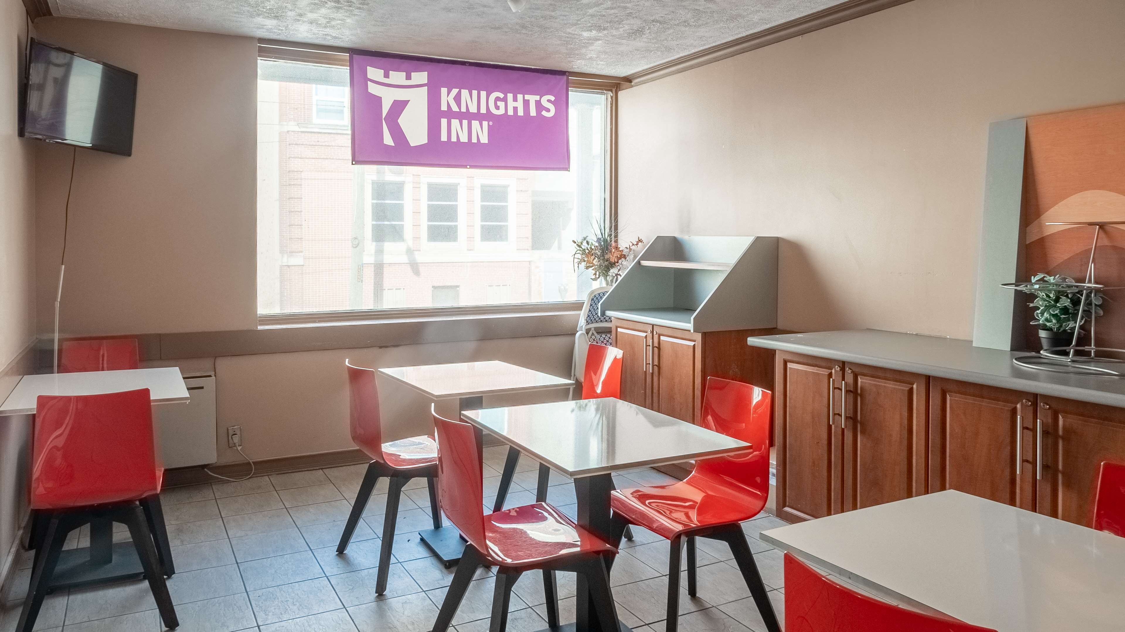A hotel room with a dining area featuring red chairs and white tables, a kitchen area, and a large window with a view of buildings outside.