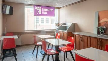 A hotel room with a dining area featuring red chairs and white tables, a kitchen area, and a large window with a view of buildings outside.