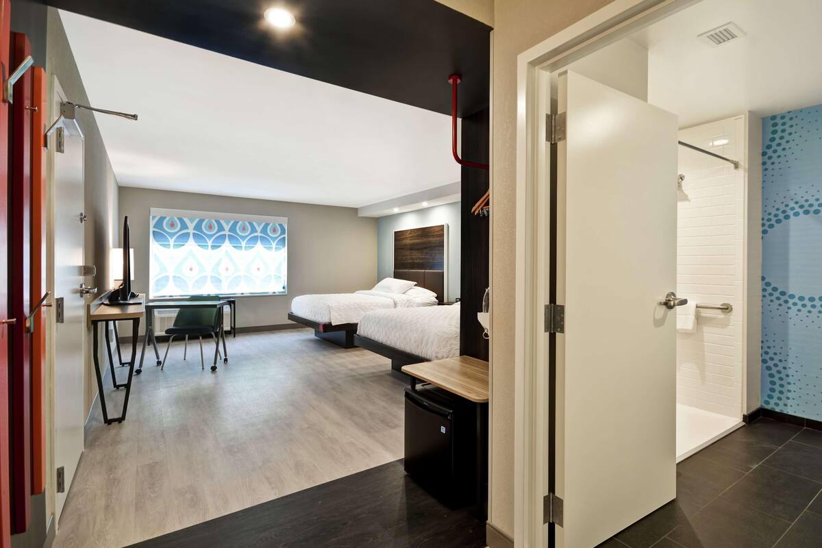 Habitación de hotel moderna con cama, escritorio, silla, una kitchenette pequeña y un baño con ducha e inodoro.