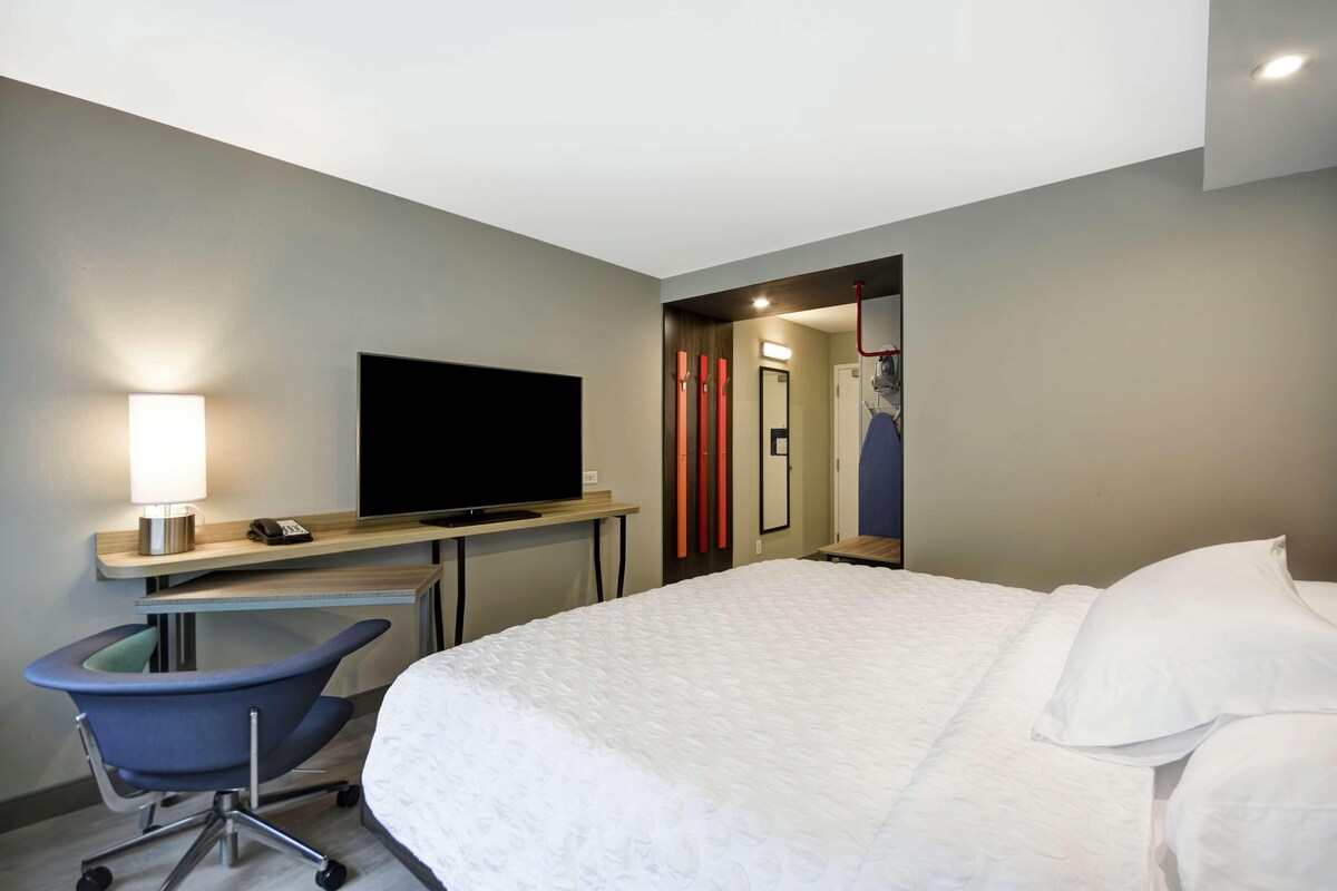 A hotel room with a bed, a desk with a TV, a chair, and a bathroom visible through an open door.