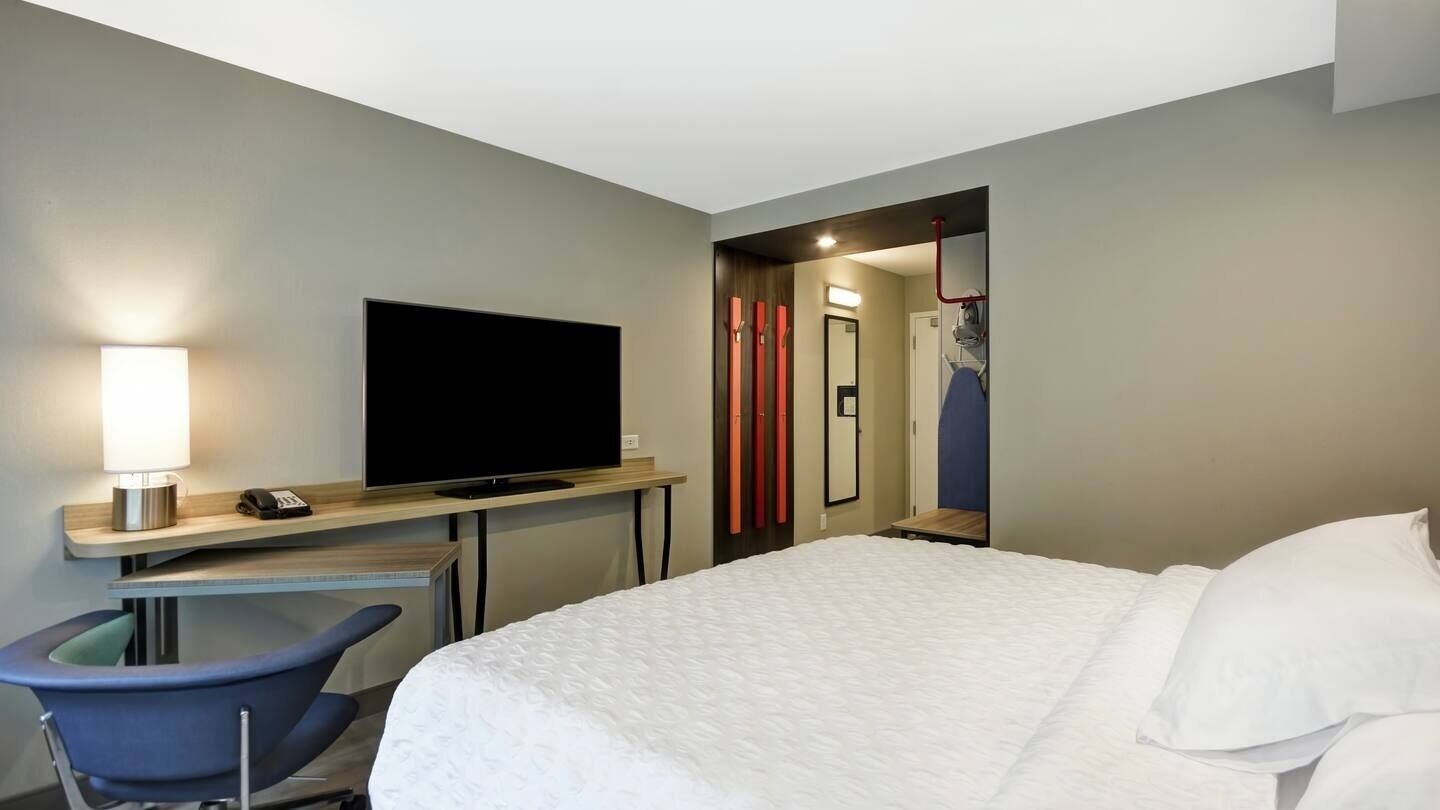 Habitación de hotel con cama, escritorio con televisor, silla y baño visible a través de una puerta abierta.