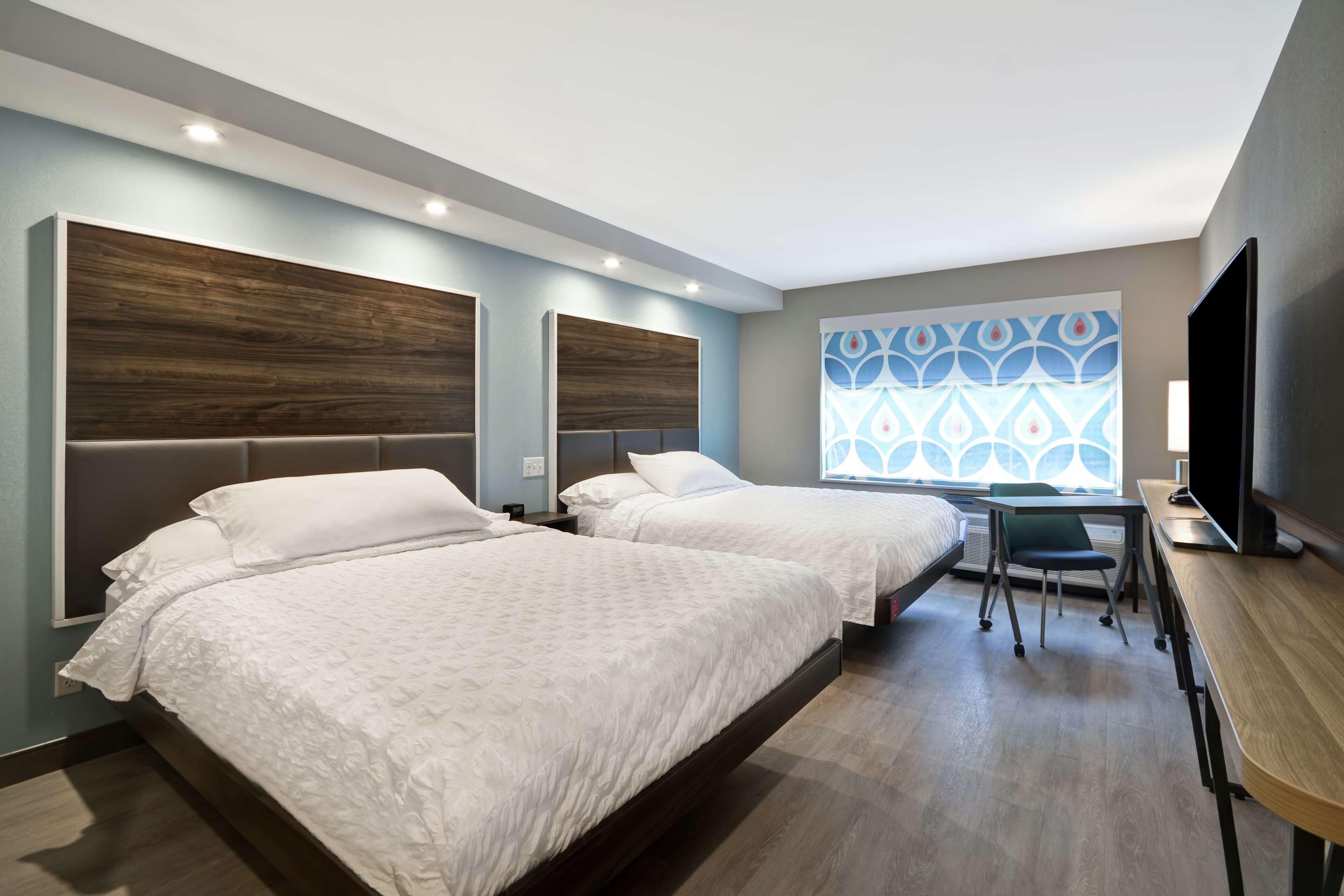 Habitación de hotel con dos camas, un escritorio, una silla, un televisor y un ventanal con un diseño geométrico.