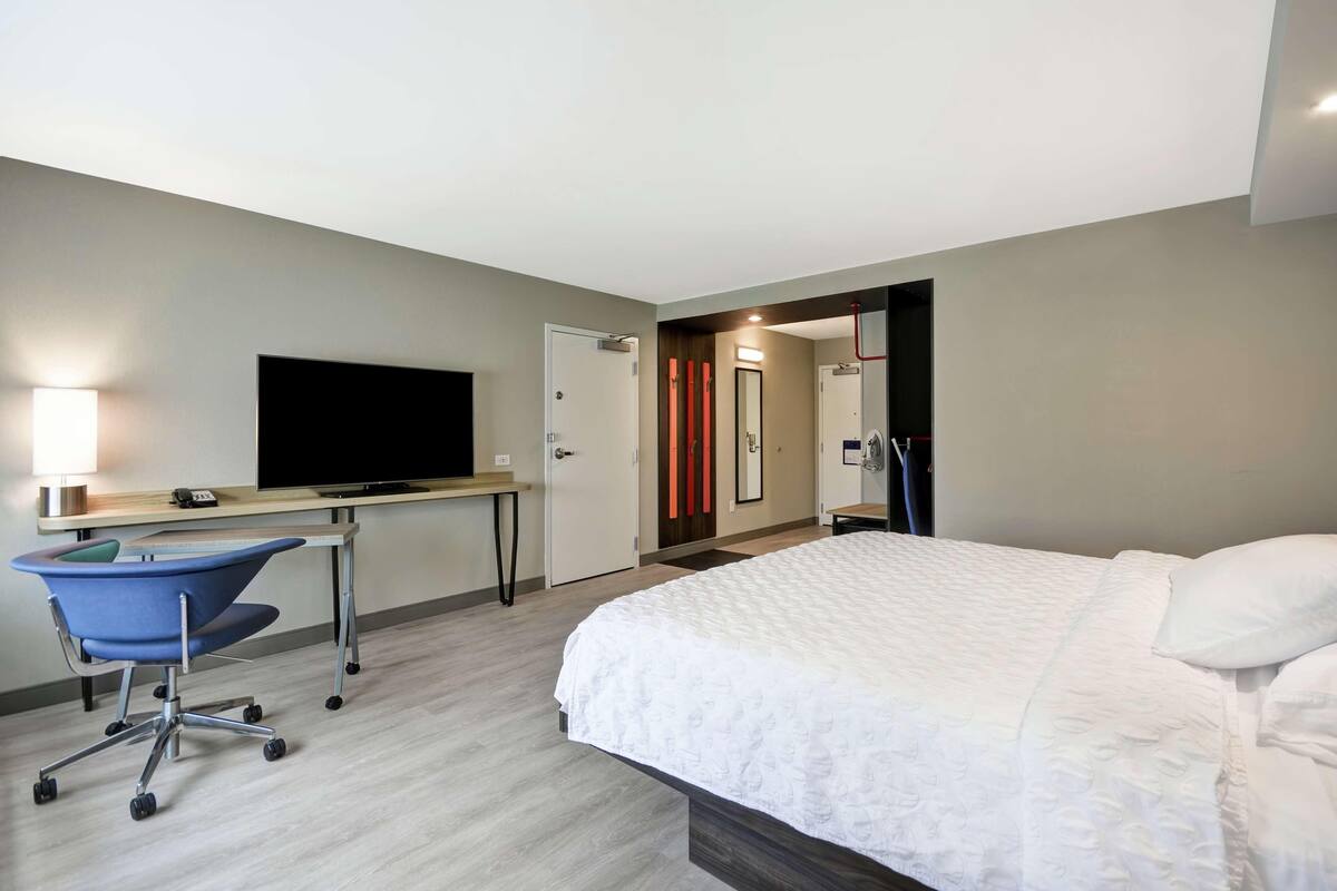 Una habitación de hotel con cama, escritorio, silla, televisor y espejo.