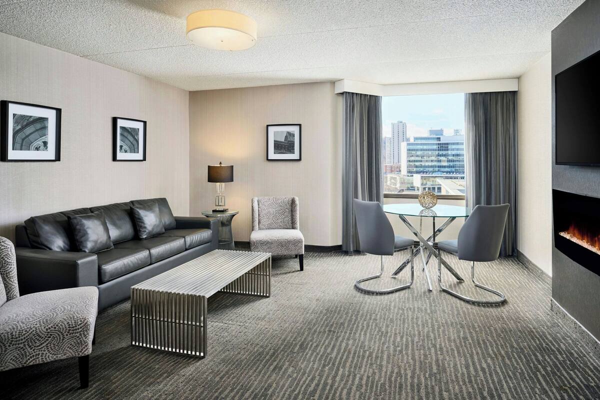 A modern hotel room with a black leather sofa, a patterned armchair, a glass coffee table, and a fireplace.