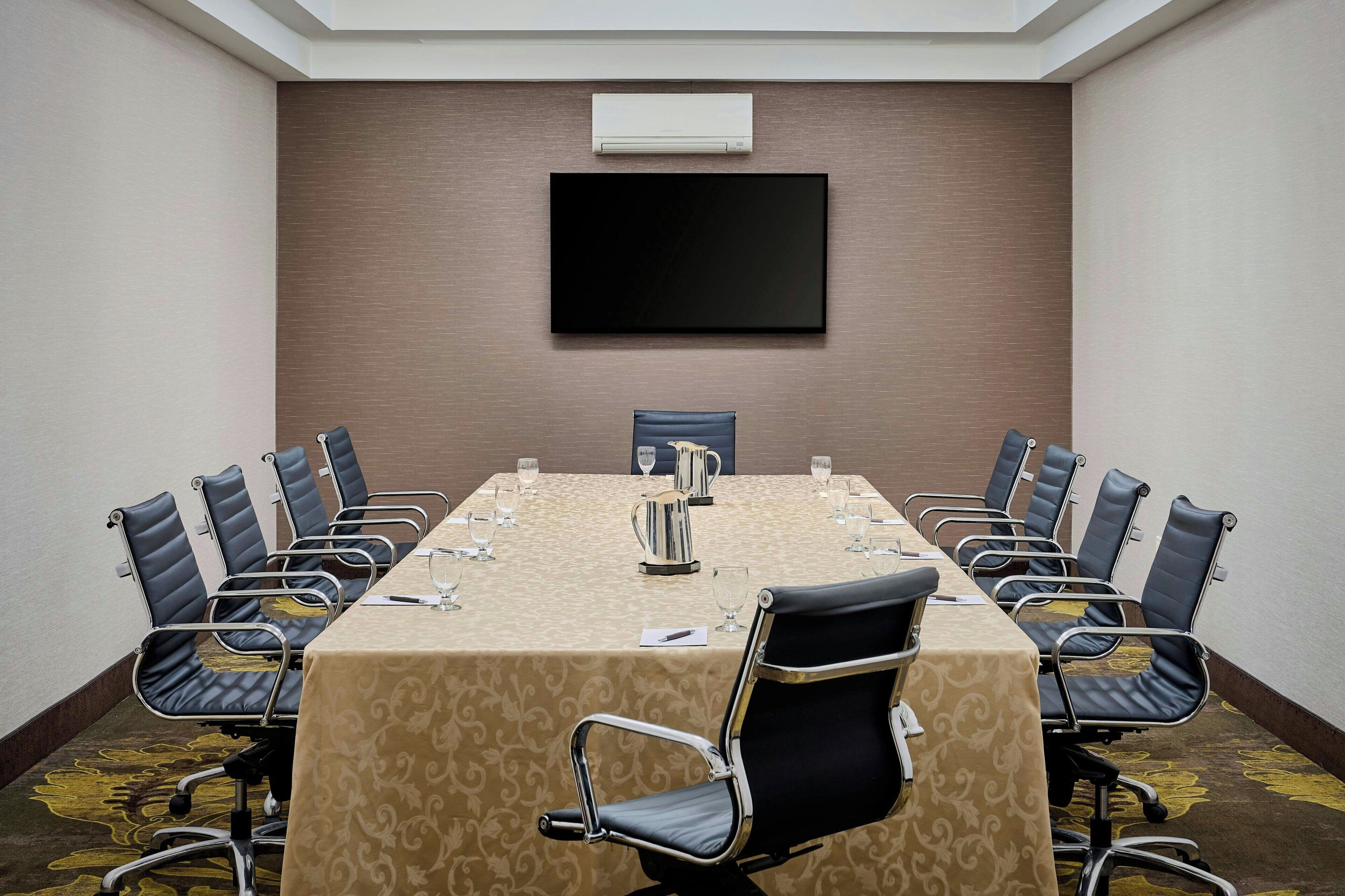 A conference room with a long table, black leather chairs, a flat-screen TV, and an air conditioning unit on the wall.
