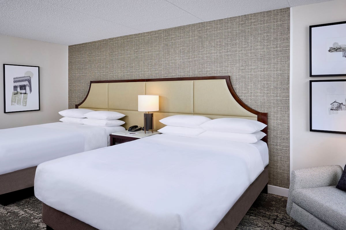 A hotel room with two beds, a nightstand with a lamp, and a chair.
