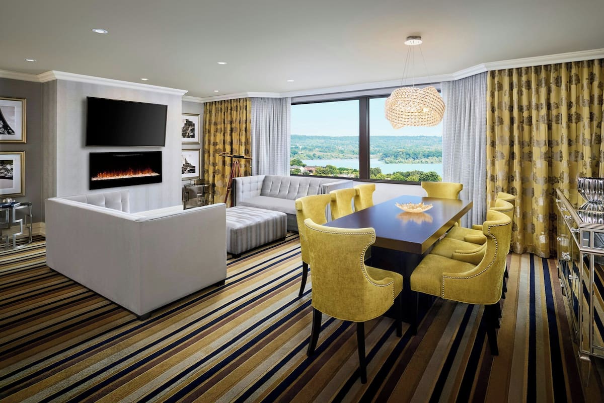 A modern hotel room with a flat-screen TV, a fireplace, a sofa, a dining table with yellow chairs, and a large window with a view of the outdoors.