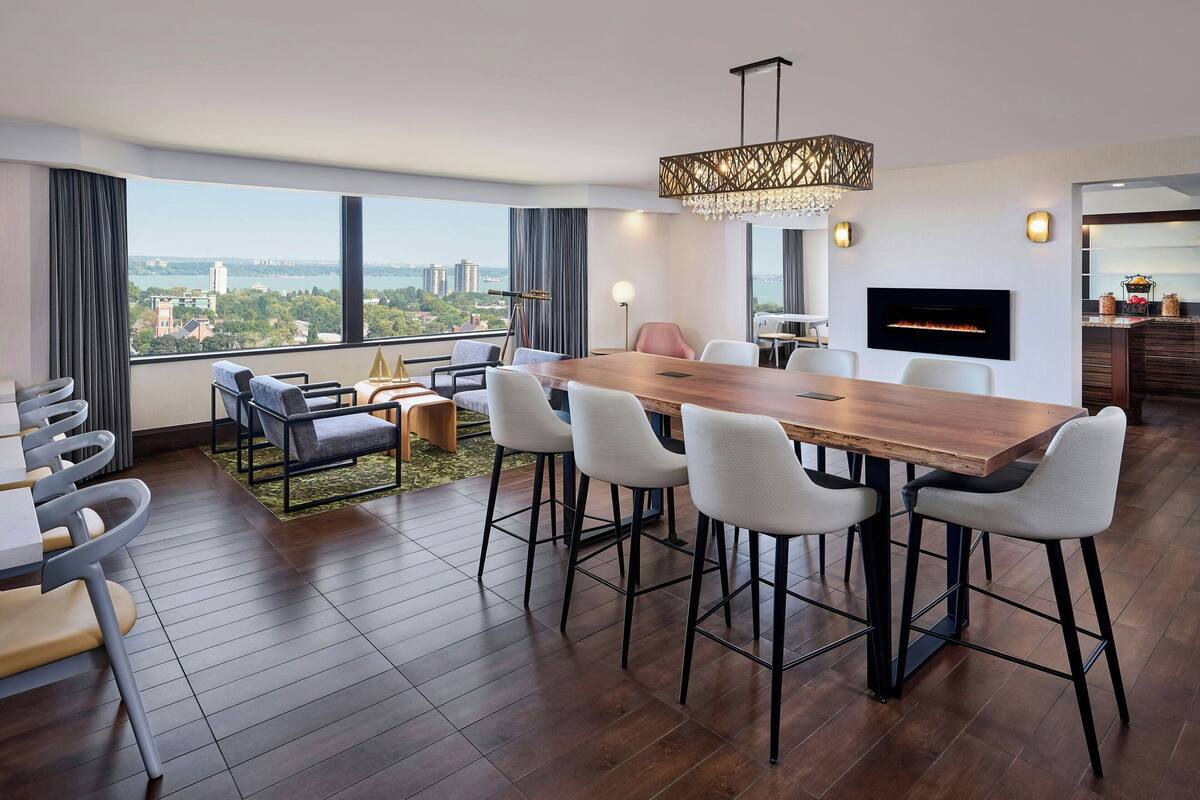 A modern hotel room with a large dining table, comfortable seating, a fireplace, and a city view.