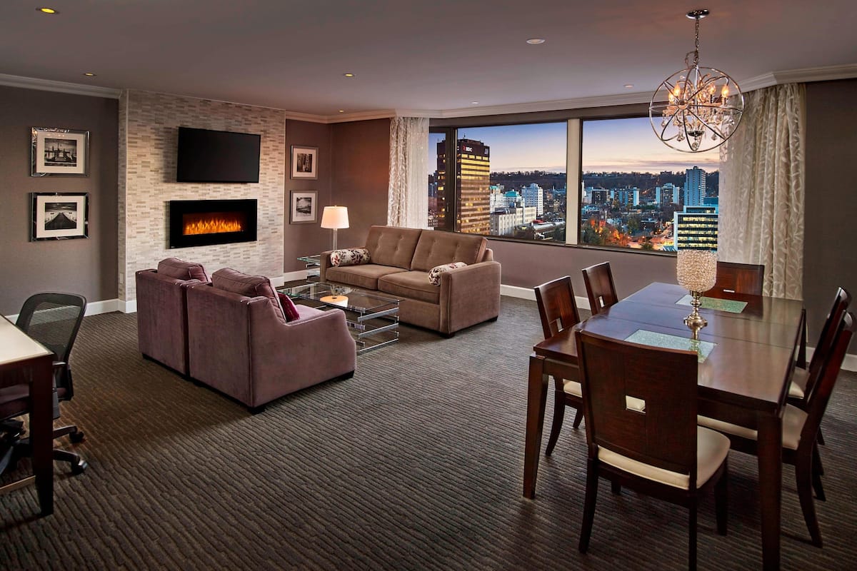 A modern hotel room with a fireplace, a large window with a city view, a flat-screen TV, and a dining area.