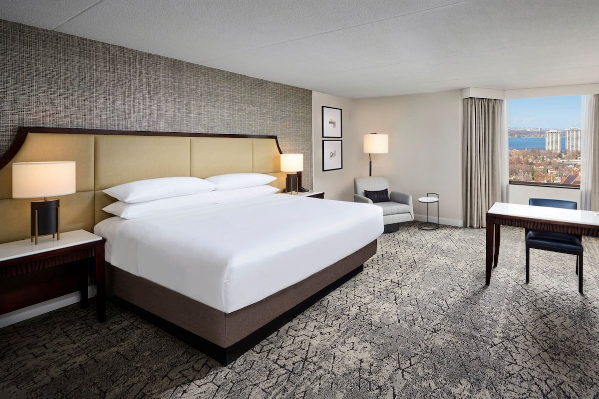 A hotel room with a large bed, two bedside tables with lamps, a small round table, a chair, and a view of the city through a large window.