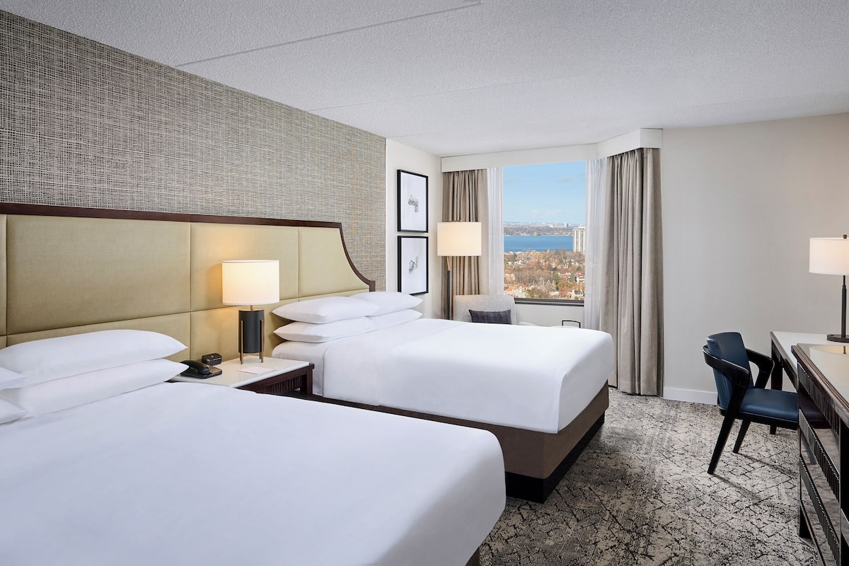 A hotel room with two beds, a desk, a chair, and a large window offering a city view.
