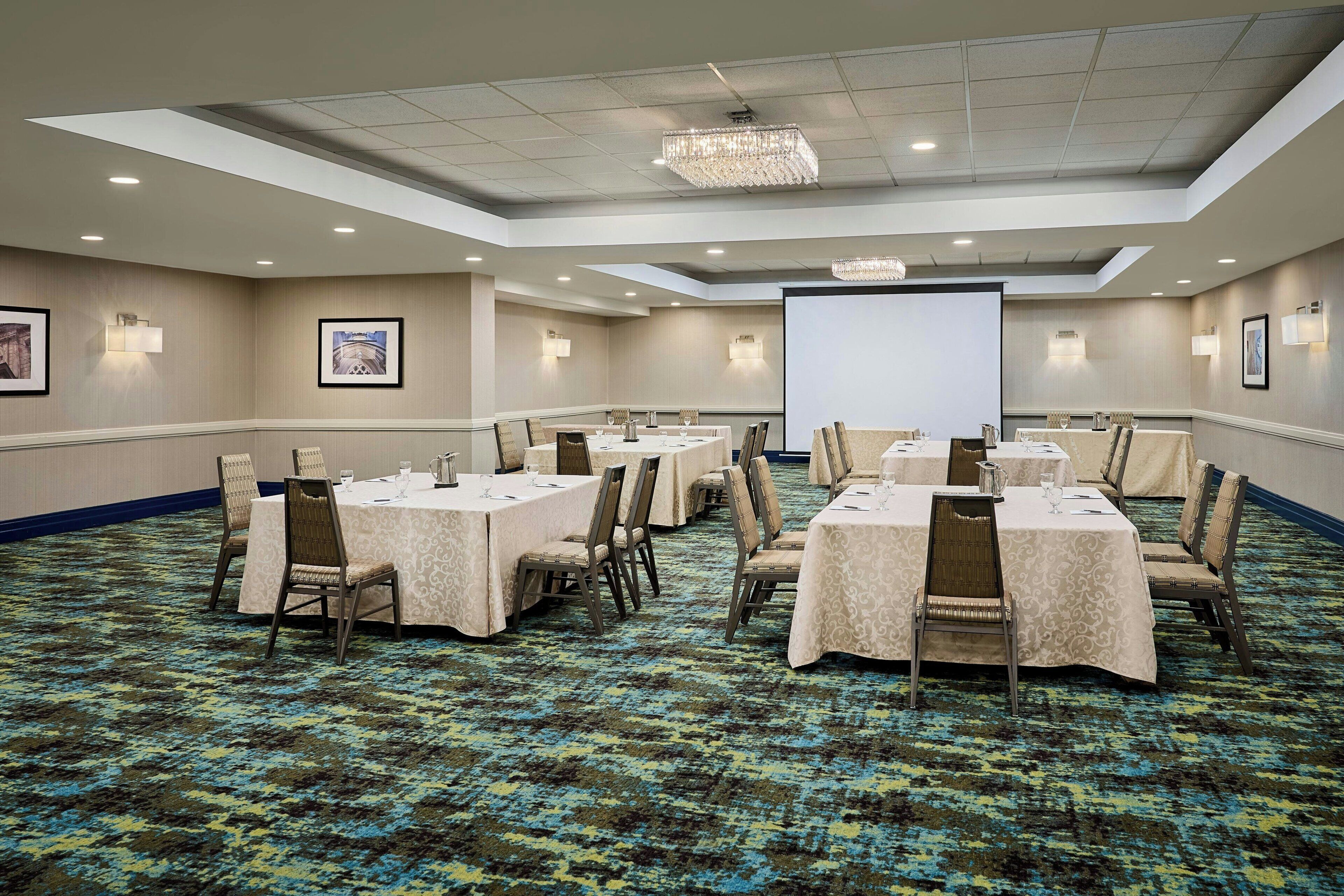 A conference room with multiple tables and chairs, a projector screen, and framed pictures on the walls.