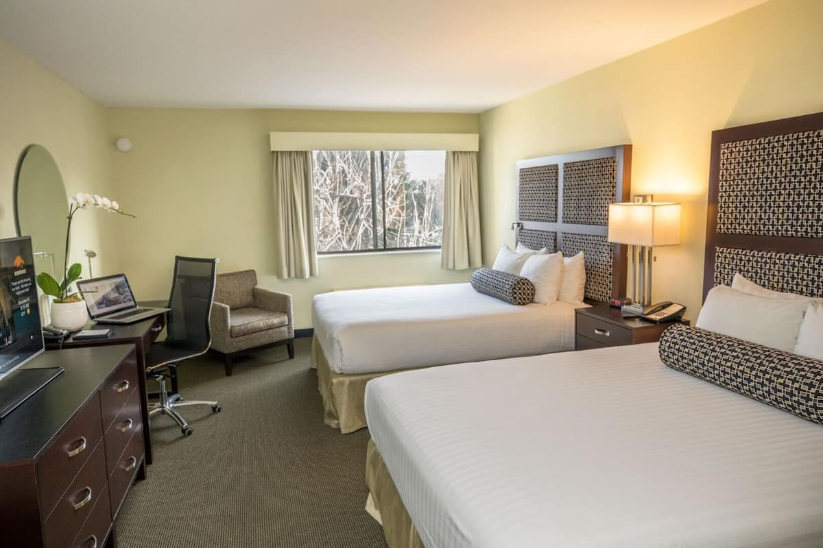 A hotel room with two beds, a desk with a laptop, a chair, a desk lamp, and a window with curtains.